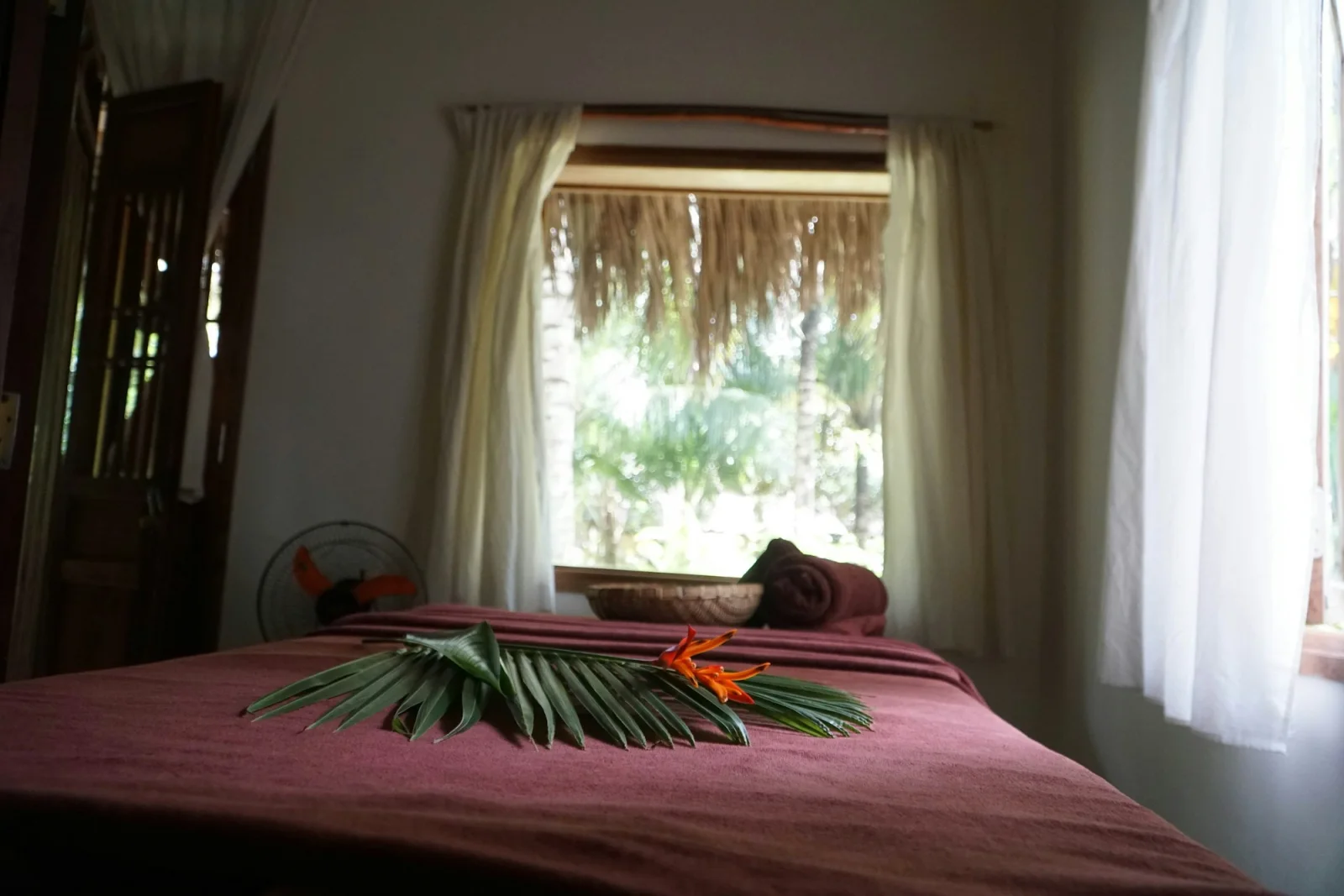 Giường trị liệu ở spa resort Cam Ranh phủ khăn đỏ trầm, lá cọ và hoa trang trí, cửa sổ nhìn ra vườn